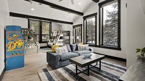 A modern living room with a sectional sofa, a coffee table, and large windows offering a view of trees. - Pet-Friendly Tahoe Escape | Hot Tub | King Suite | Arcade | Near Trails & Lake (Olympic Valley)