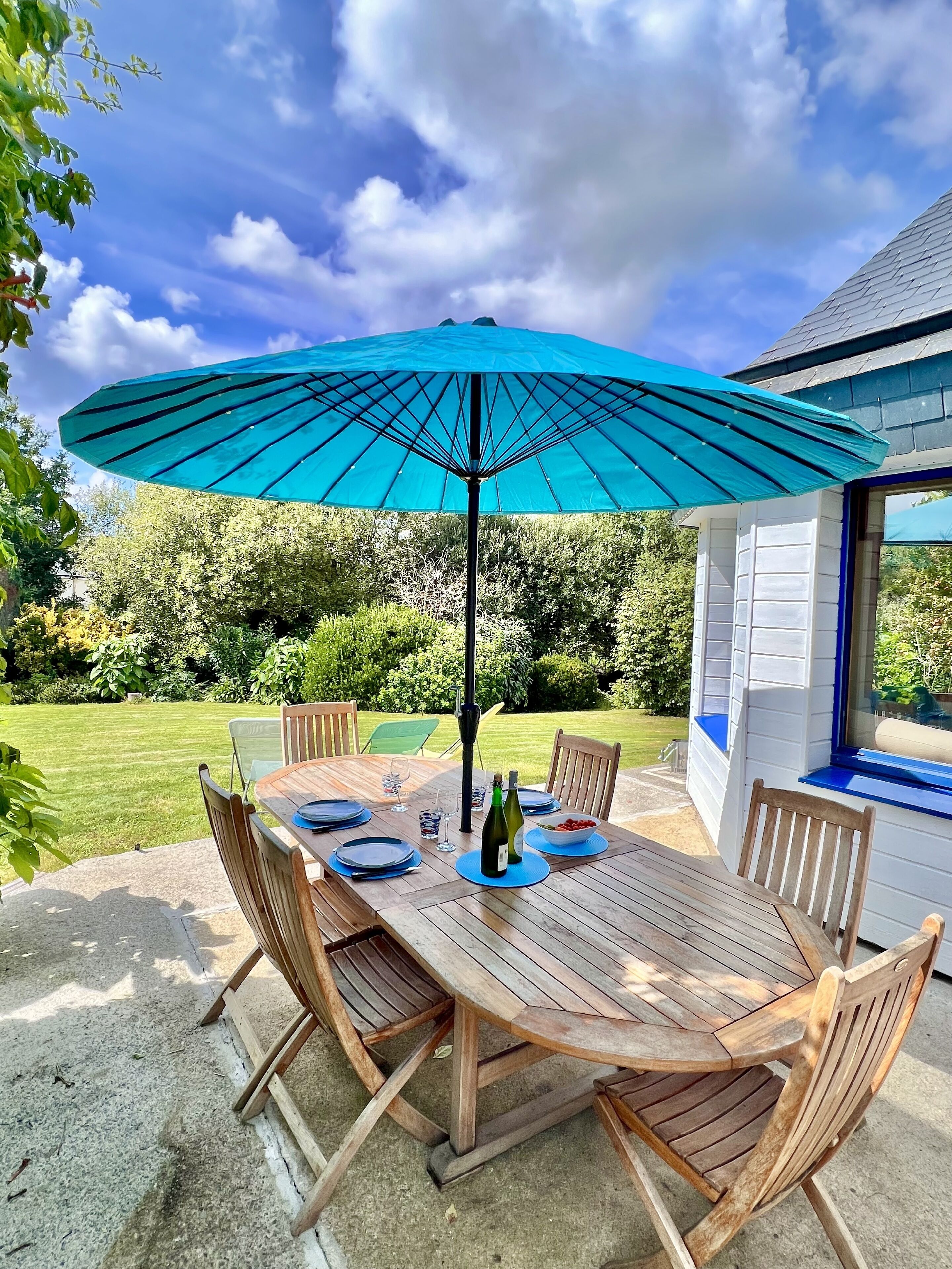 A wooden table set outdoors with a blue umbrella, surrounded by chairs, and a garden in the background.
