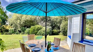 Una mesa de madera al aire libre con un paraguas azul, rodeada de sillas, y un jardín al fondo.