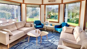 Un solárium con techo alto, ventanas grandes, un sofá color crema, dos sillones azules y una mesita con vasos.