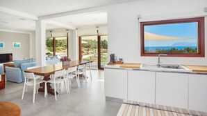 Een moderne keuken met een groot raam, witte kasten en een eethoek met een houten tafel en witte stoelen.
