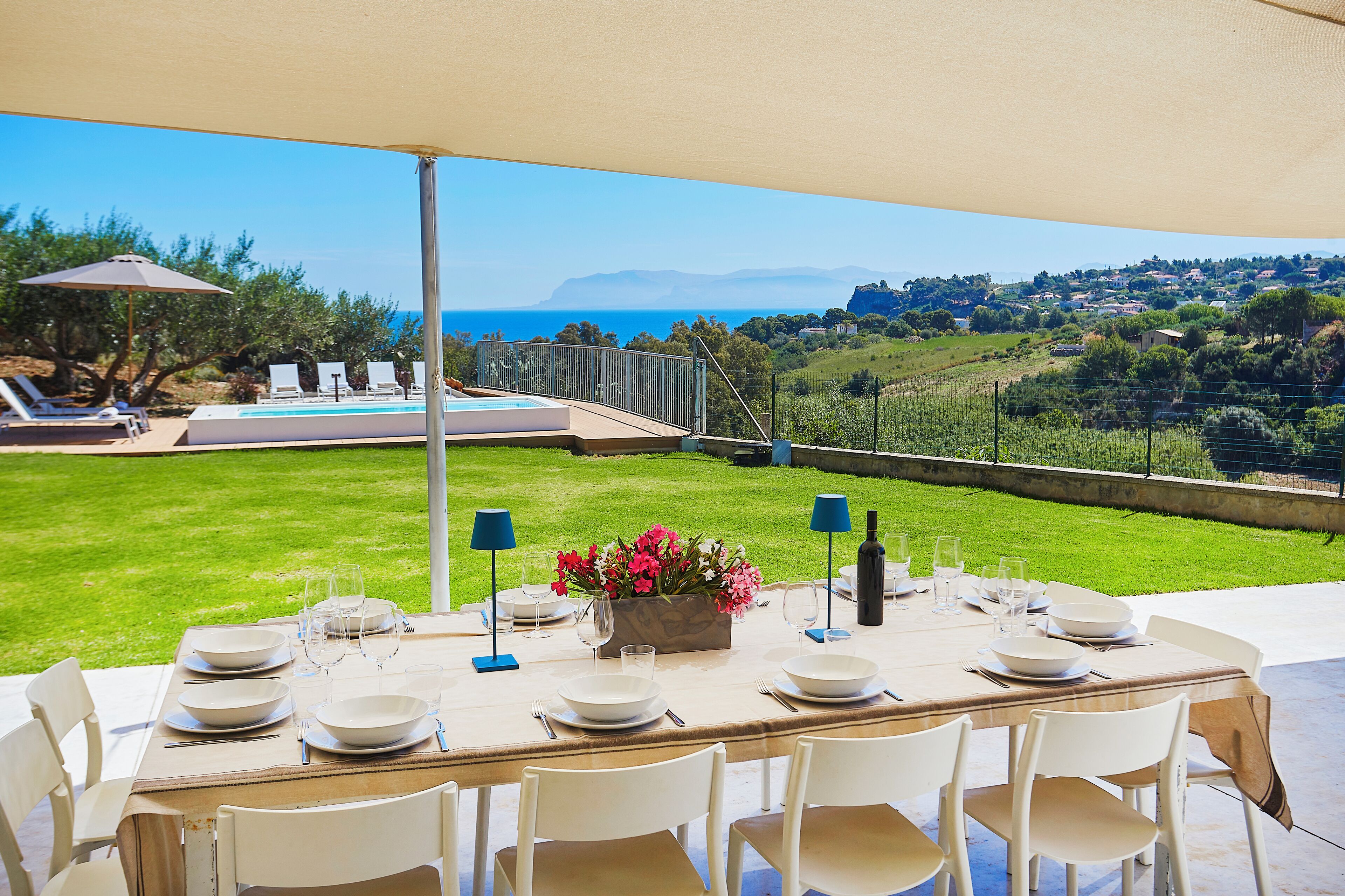 A table set for dining with a view of the sea and a grassy area with lounge chairs.
