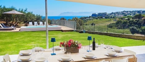 A table set for dining with a view of the sea and a grassy area with lounge chairs.