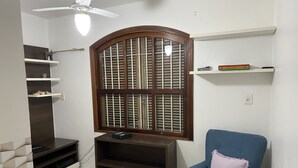 A room with a built-in entertainment unit, a blue armchair with a pink cushion, a ceiling fan, and a large window with wooden shutters. - Sobrado Tranquilo, rua Silenciosa (Jardim São Paulo São Paulo)