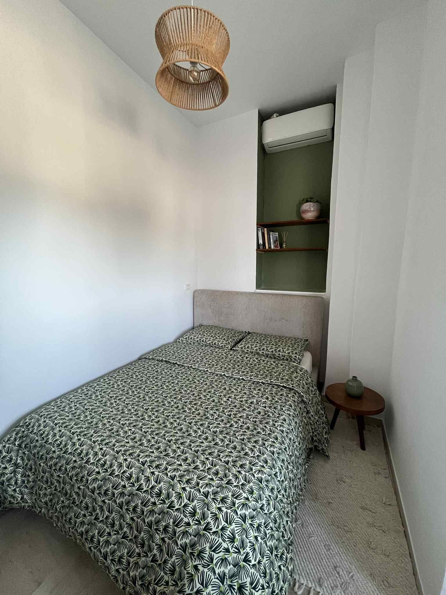 A small, neatly arranged bedroom with a single bed, a small table, a hanging light fixture, and a built-in shelf with books and decorative items.