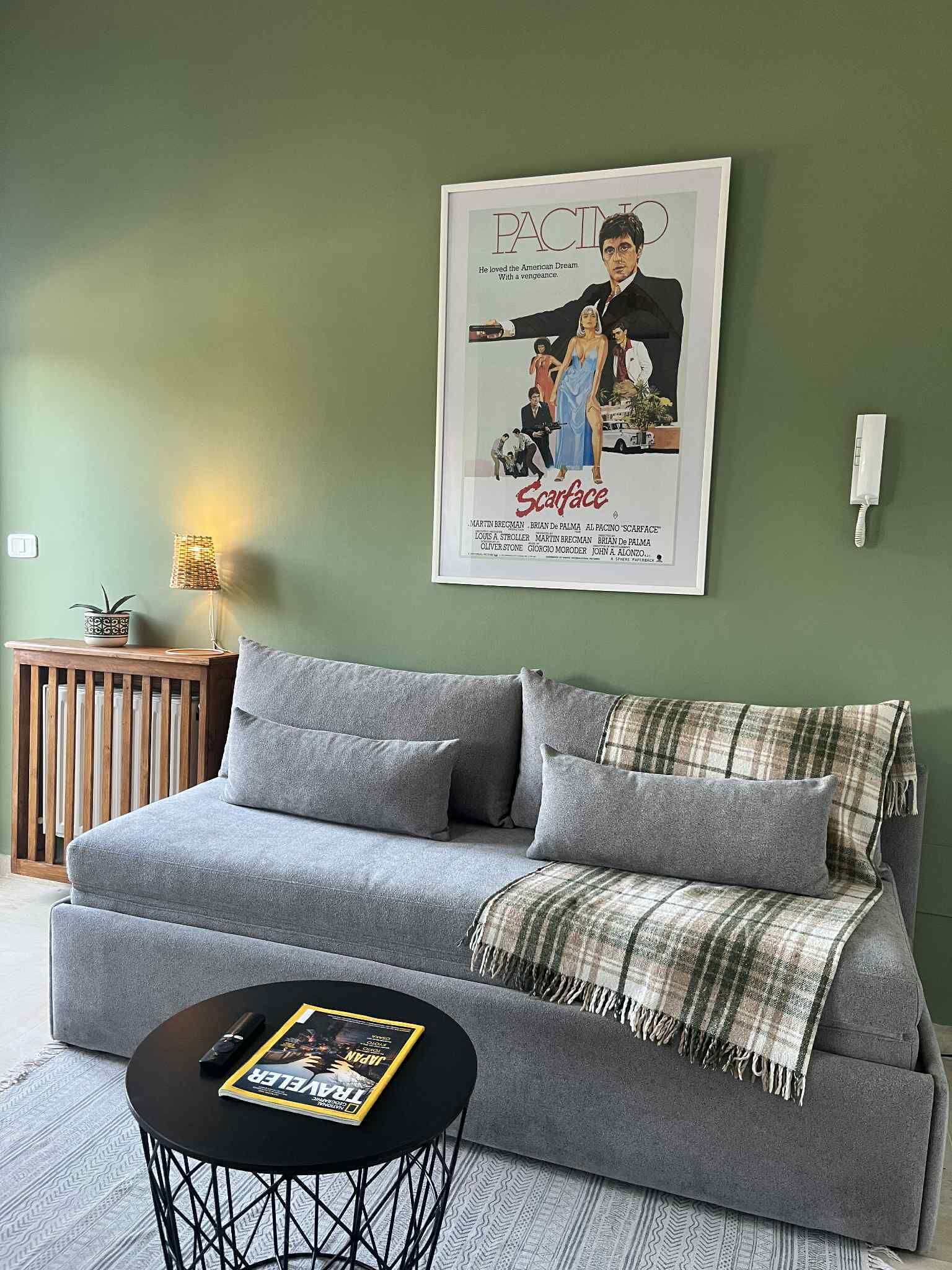 A living room with a grey sofa, a plaid throw blanket, a magazine on a small round table, and a framed movie poster on the wall.