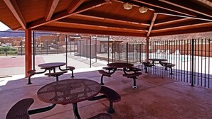 A covered outdoor seating area with metal tables and benches, a chain-link fence, and a view of a parking lot and buildings in the background. - The Howling Coyote - Moab's Adventure Basecamp (Moab)