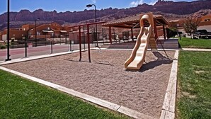 A playground with a slide and swings, surrounded by a fenced area with a mountain backdrop. - The Howling Coyote - Moab's Adventure Basecamp (Moab)