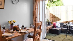 A dining area with a wooden table set for two, a clock on the wall, and a view of the backyard through a sliding door. - 2 bedroom accommodation in Mundesley (Mundesley)