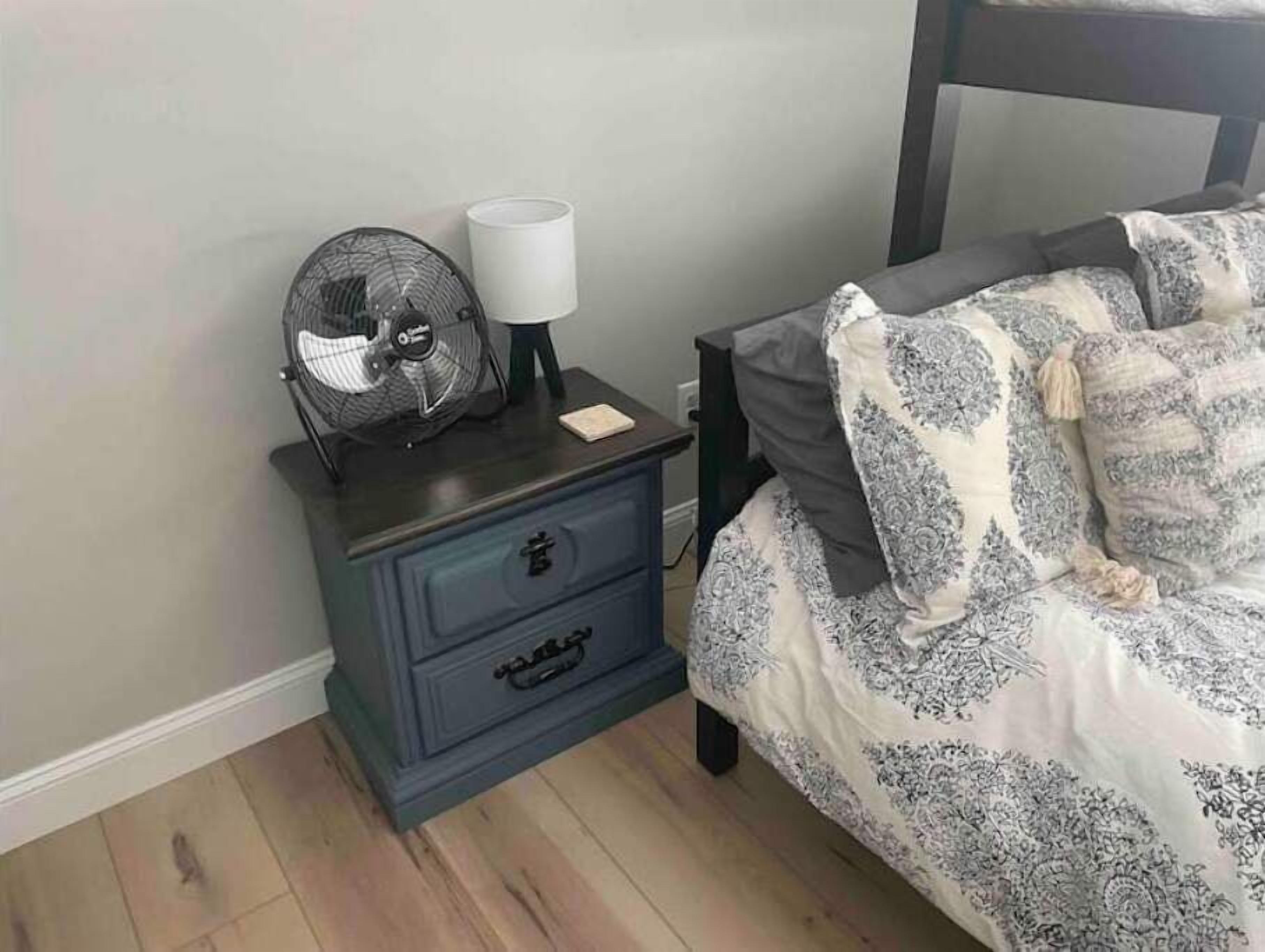 A bedroom with a dark wooden nightstand, a white lamp, a black fan, and a bed with patterned bedding.