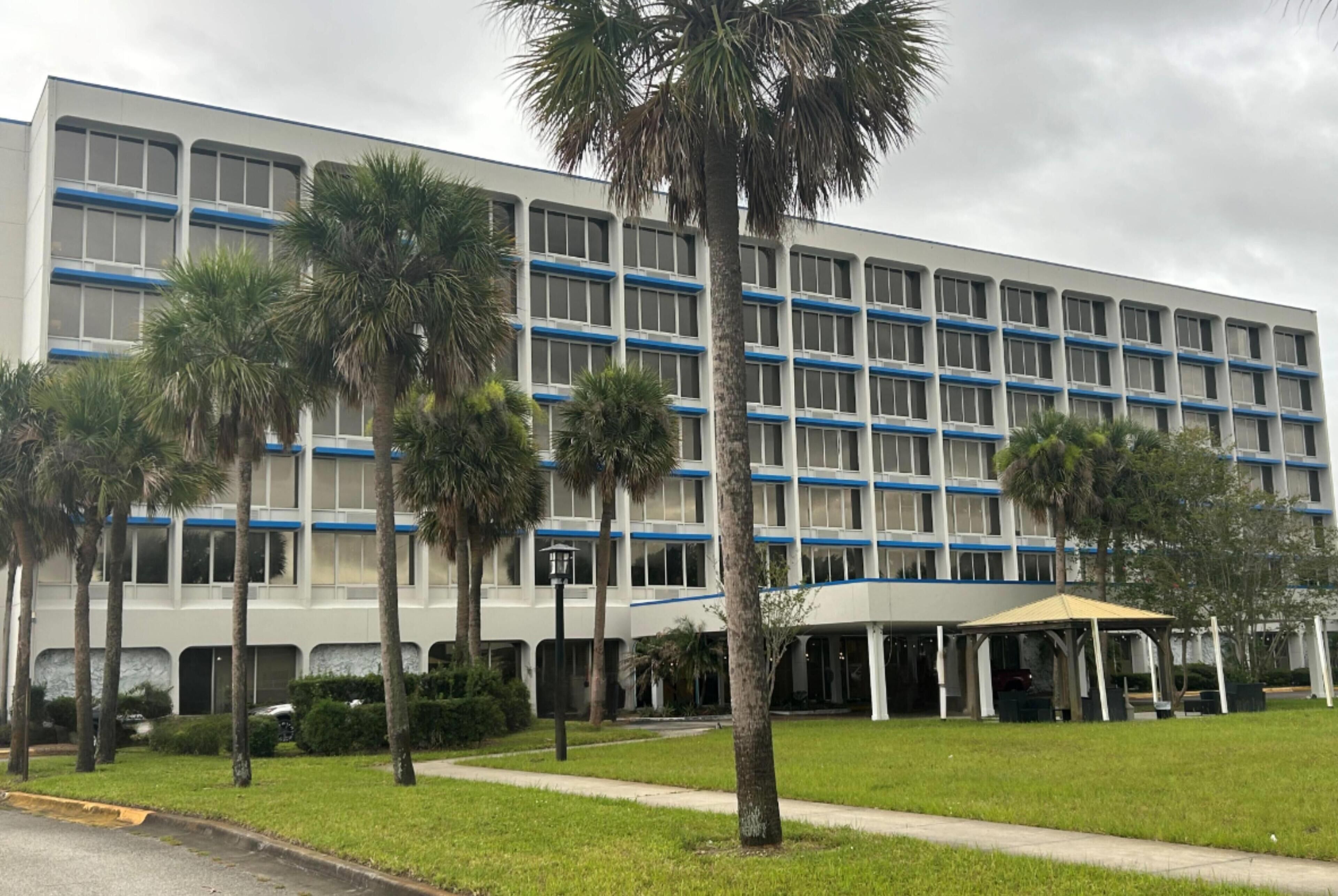 Un edificio de varios pisos con techo plano, rodeado de palmeras y una zona de estar al aire libre techada.