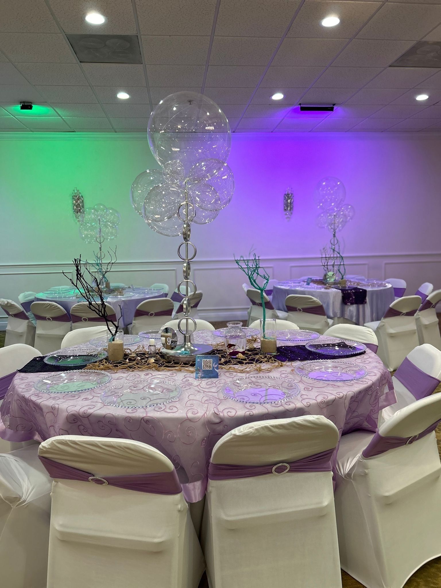 Une salle de banquet avec des tables rondes recouvertes de nappes violettes, des chaises blanches ornées de rubans violets, et des éléments décoratifs tels que des ornements en verre et des branches.