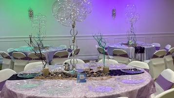 Une salle de banquet avec des tables rondes recouvertes de nappes violettes, des chaises blanches ornées de rubans violets, et des éléments décoratifs tels que des ornements en verre et des branches.