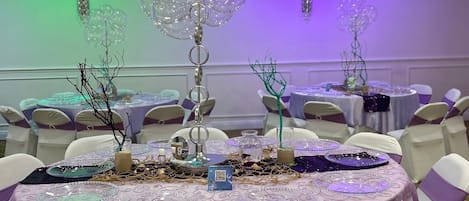 A banquet hall with round tables covered in purple tablecloths, white chairs with purple sashes, and decorative elements like glass ornaments and branches.