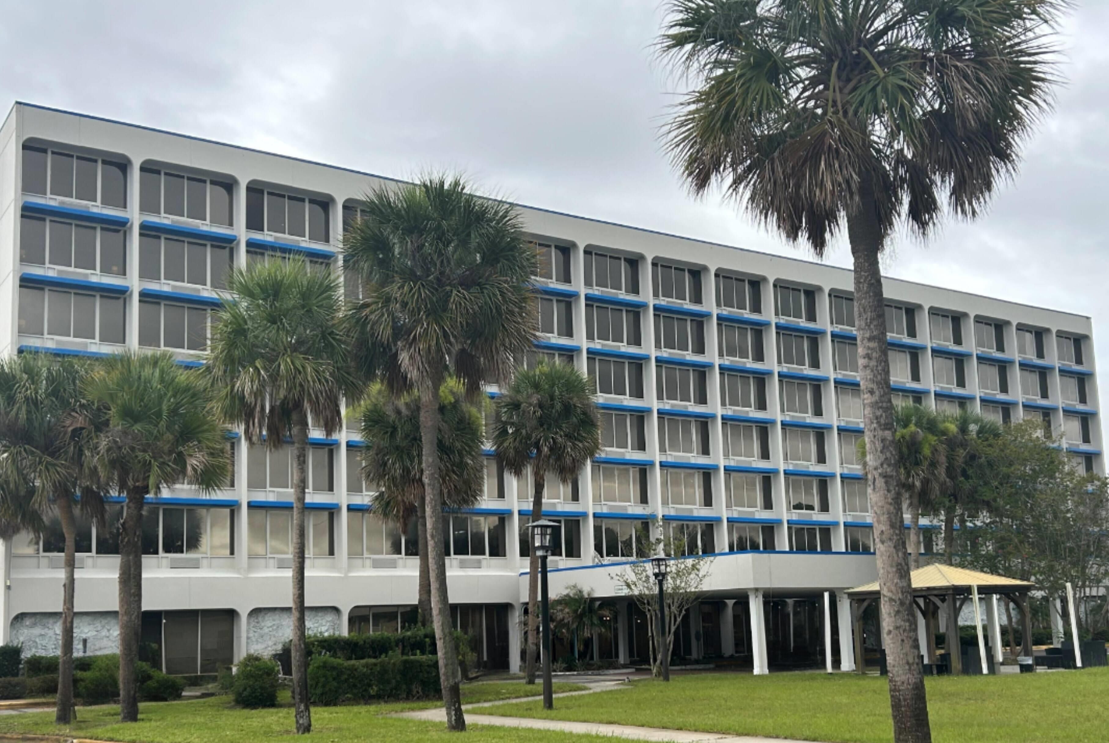 A multi-story building with a white facade and blue accents, surrounded by palm trees and a covered walkway.