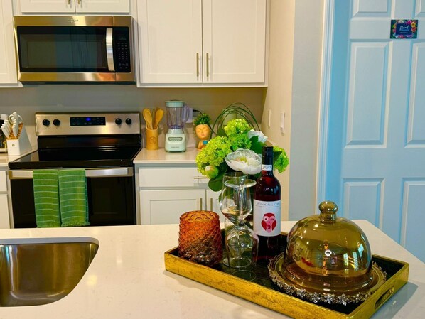 A kitchen with white cabinets, a stainless steel microwave, a stove, and a sink. There is a tray with a bottle of Winking Owl wine, a glass, and a decorative item.