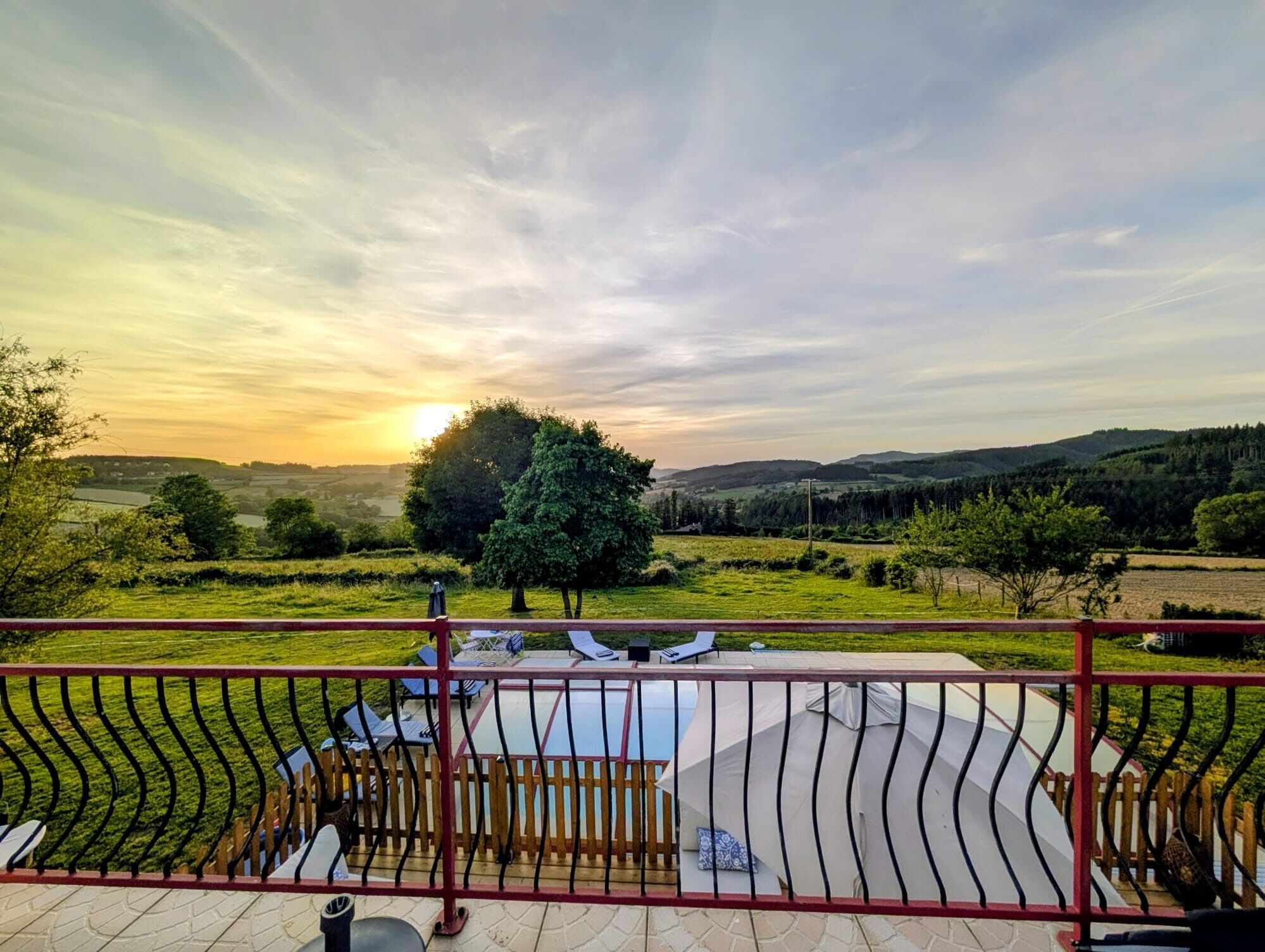 Une terrasse offrant une vue sur un coucher de soleil se déclinant sur un paysage vallonné, avec un arbre et un espace clos aménagé avec du mobilier d’extérieur.