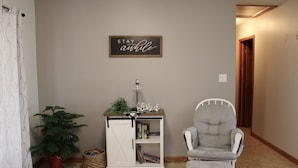 A hallway with a white chair, a small white cabinet with books, a potted plant, and a sign that says "STAY awhile".