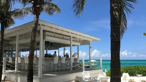 A covered outdoor seating area with white chairs and tables, overlooking a beach with clear turquoise water and palm trees. - Delaporte Point Near waterpark Private Beach! (Nassau)