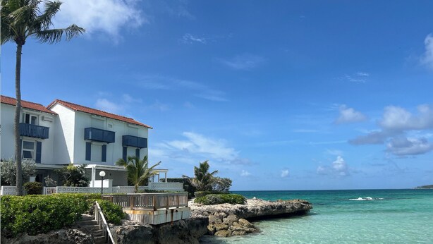 A beachfront property with a white building, clear turquoise water, and a rocky shoreline. - Delaporte Point Near waterpark Private Beach! (Nassau)