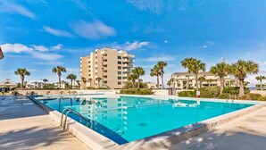 A swimming pool with a clear blue water, surrounded by palm trees and a multi-story building in the background. - Pensacola Beach Waterfront Condo with Relaxing View. Family Friendly! (Pensacola Beach)