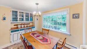 A dining area with a wooden table, chairs, a chandelier, and a window with a view of greenery. - 3BR- Walk or Bike to Acadia! [Carpenters Quarters] (Mount Desert)