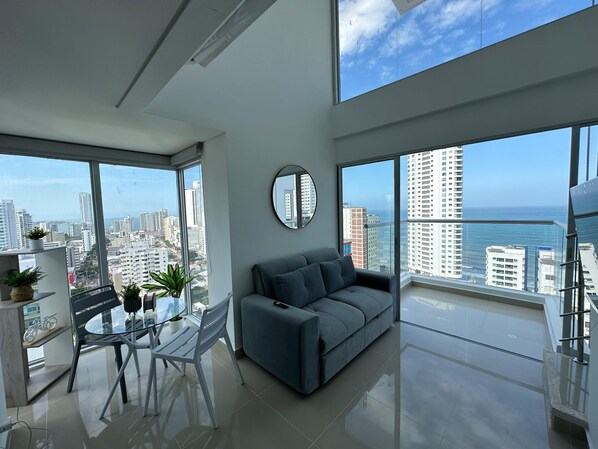 A modern living room with a large window, a grey sofa, a round mirror, and a dining area with a glass table and chairs.