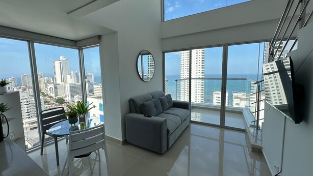 A modern living room with a grey sofa, a round mirror, and a dining area with a table and chairs. Large windows offer a view of the city and the ocean. - Apartamento loft de 1hab vista al mar (Cartagena)