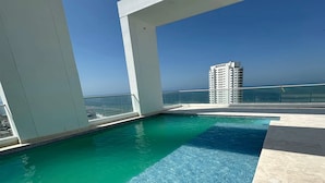 A rooftop pool with clear blue water, surrounded by white pillars and a view of a high-rise building across the water. - Apartamento loft de 1hab vista al mar (Cartagena)