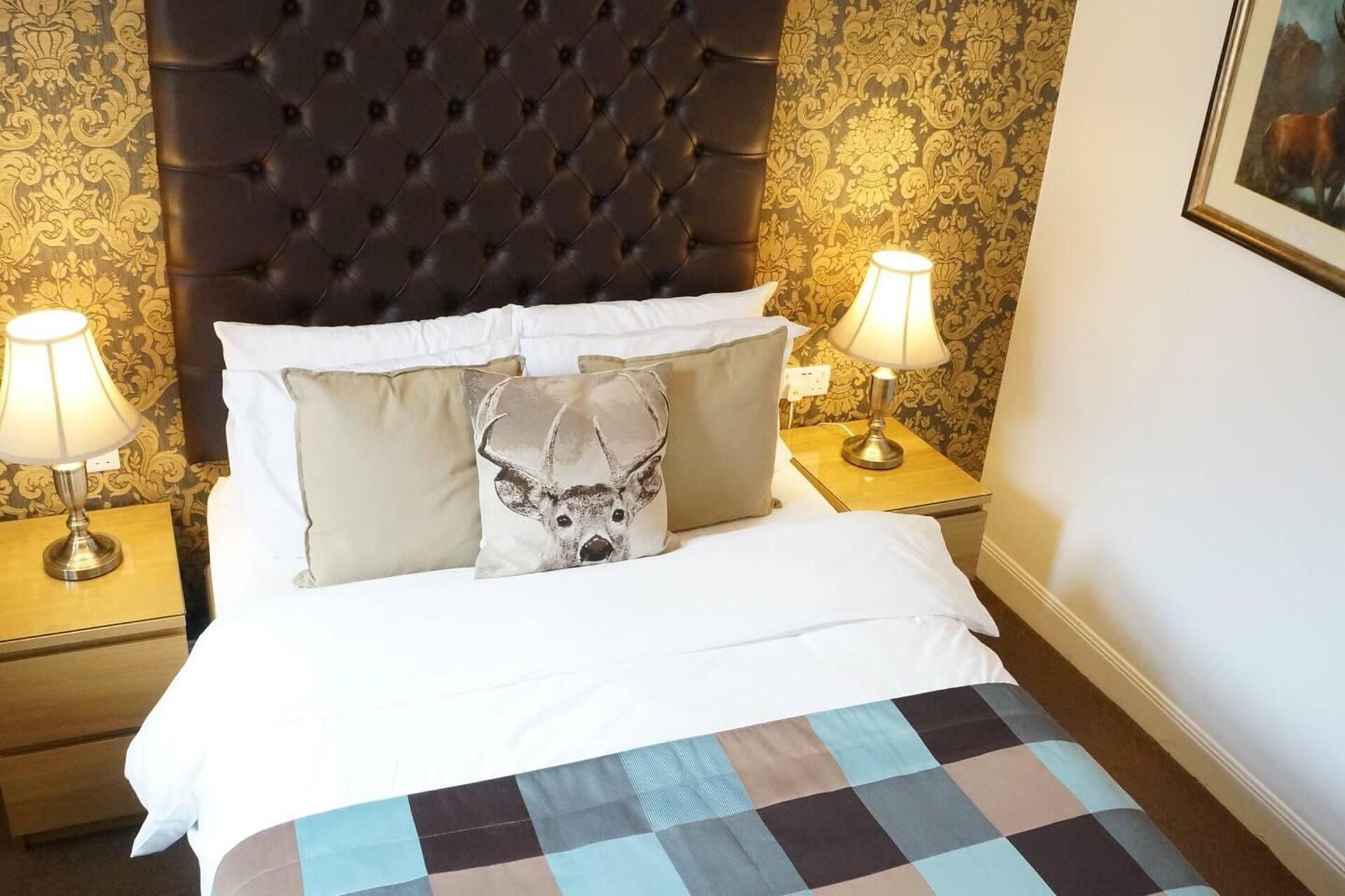 A hotel room with a tufted headboard, two bedside lamps, a patterned wall, and a framed picture on the wall.