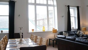 A room with a dining table set for four, a black leather sofa, two lamps, and large arched windows with curtains. - Rothesay - Apartment - Sleeps9 - Petsok - Parking (Rothesay)