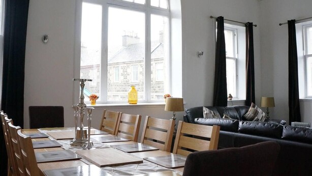 A room with a dining table set for four, a black leather sofa, and large arched windows with curtains. - Rothesay - Apartment - Sleeps9 - Petsok - Parking (Rothesay)