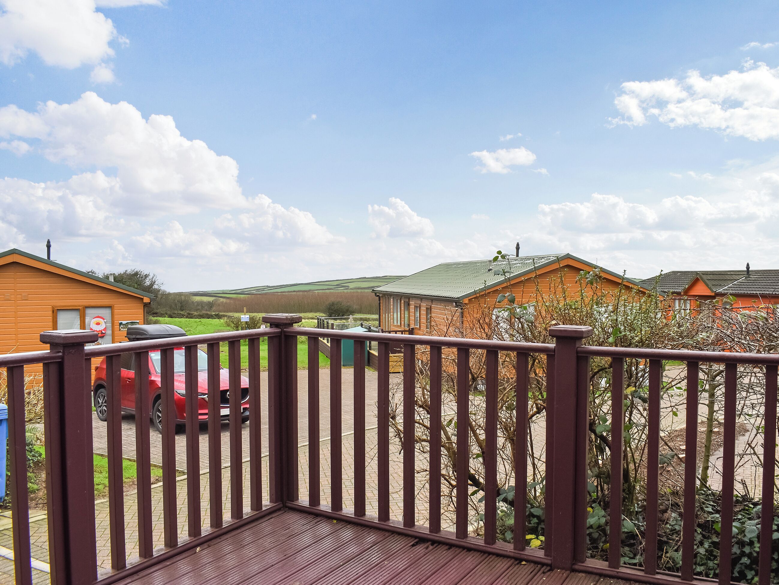 A wooden deck with a view of a red car parked in a driveway, surrounded by cabins with green roofs.