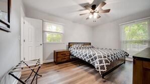 Une chambre à coucher comprenant un lit, un ventilateur de plafond, une table de chevet, une commode en bois et une fenêtre avec des stores.