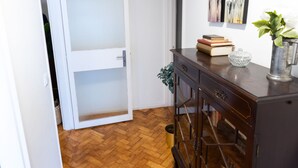 Un passadís amb un paviment de fusta en disseny de petxina, una porta blanca amb un panell de vidre mat, un armari de fusta fosca amb calaixos i una petita planta al terra.