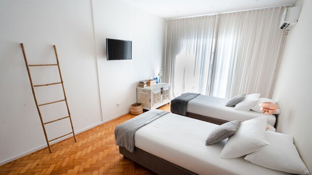 Habitació d'hotel moderna amb dos llits, un terra de fusta en disseny de petxina d'atzó, un televisor a la paret i una finestra amb cortines de tul.