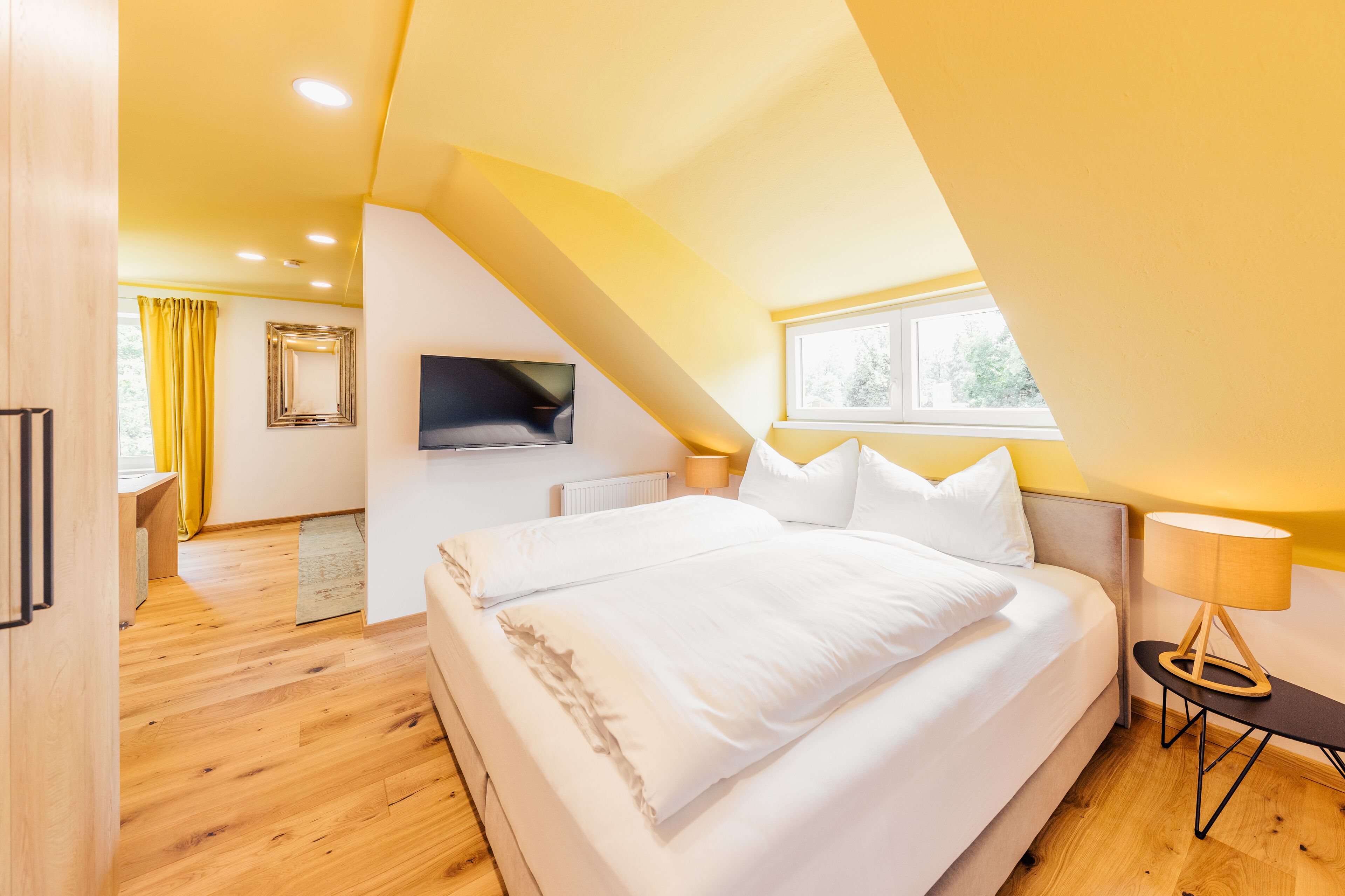 A bedroom with a large bed, a television mounted on the wall, a window, and a lamp on a side table.