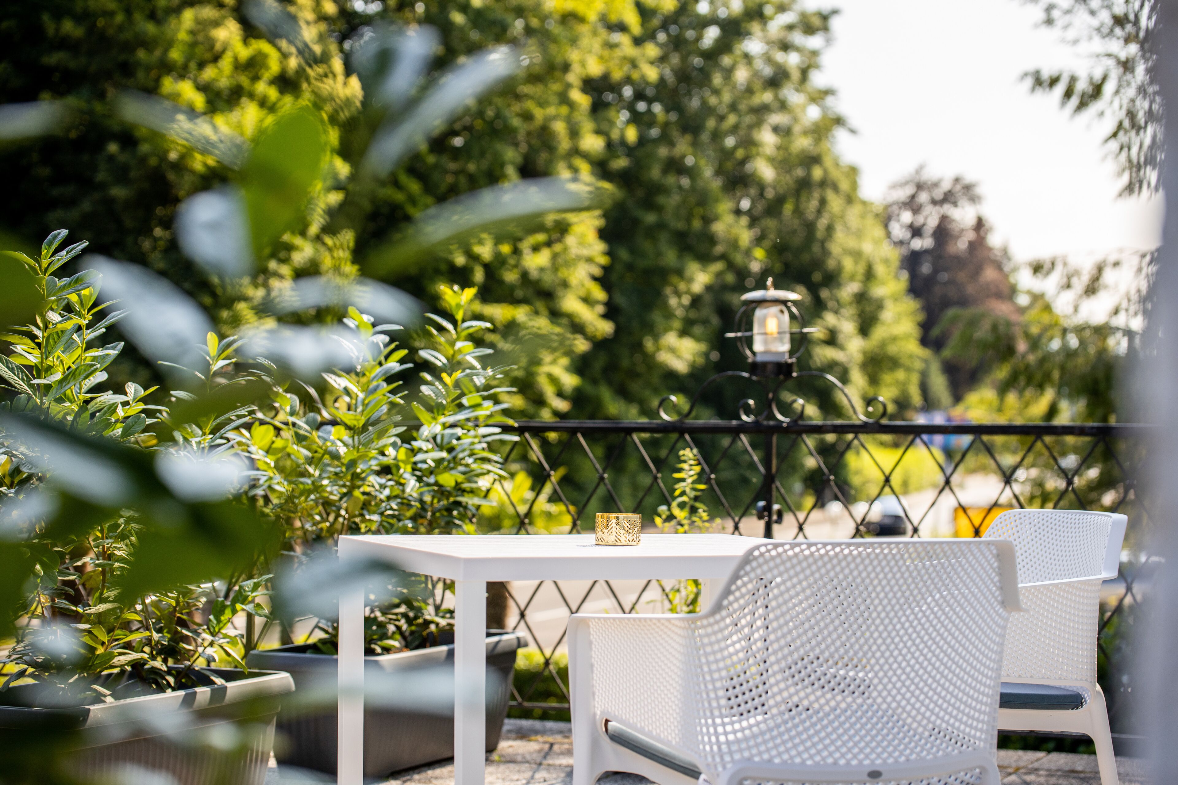 Ein weißes Tisch- und Stuhlsitzset auf einem Balkon mit Metallgeländer, umgeben von Topfpflanzen und einer Laterne.