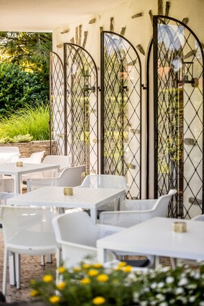 An outdoor seating area with white tables and chairs, surrounded by a metal fence. - Boutiquehotel Caravella (Velden am Wörther See)