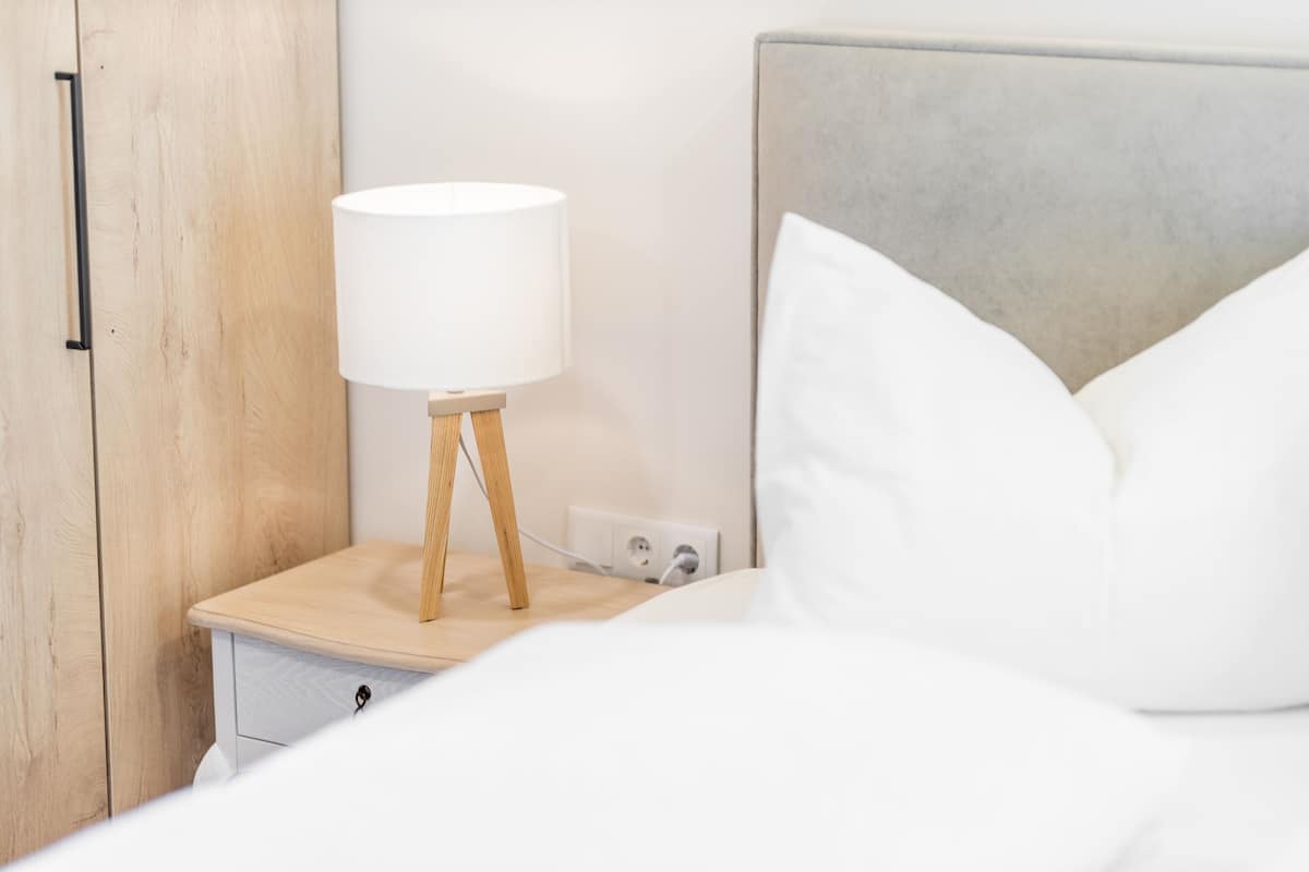 Un lit bien fait, avec du linge de lit blanc, une table de chevet en bois surmontée d’une lampe blanche et une tête de lit rembourrée de couleur grise.