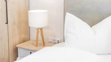 Ein ordentlich bezogenes Bett mit weißen Leinen, ein Nachttisch aus Holz mit einer weißen Lampe und ein grauer, gepolsterter Kopfteil.