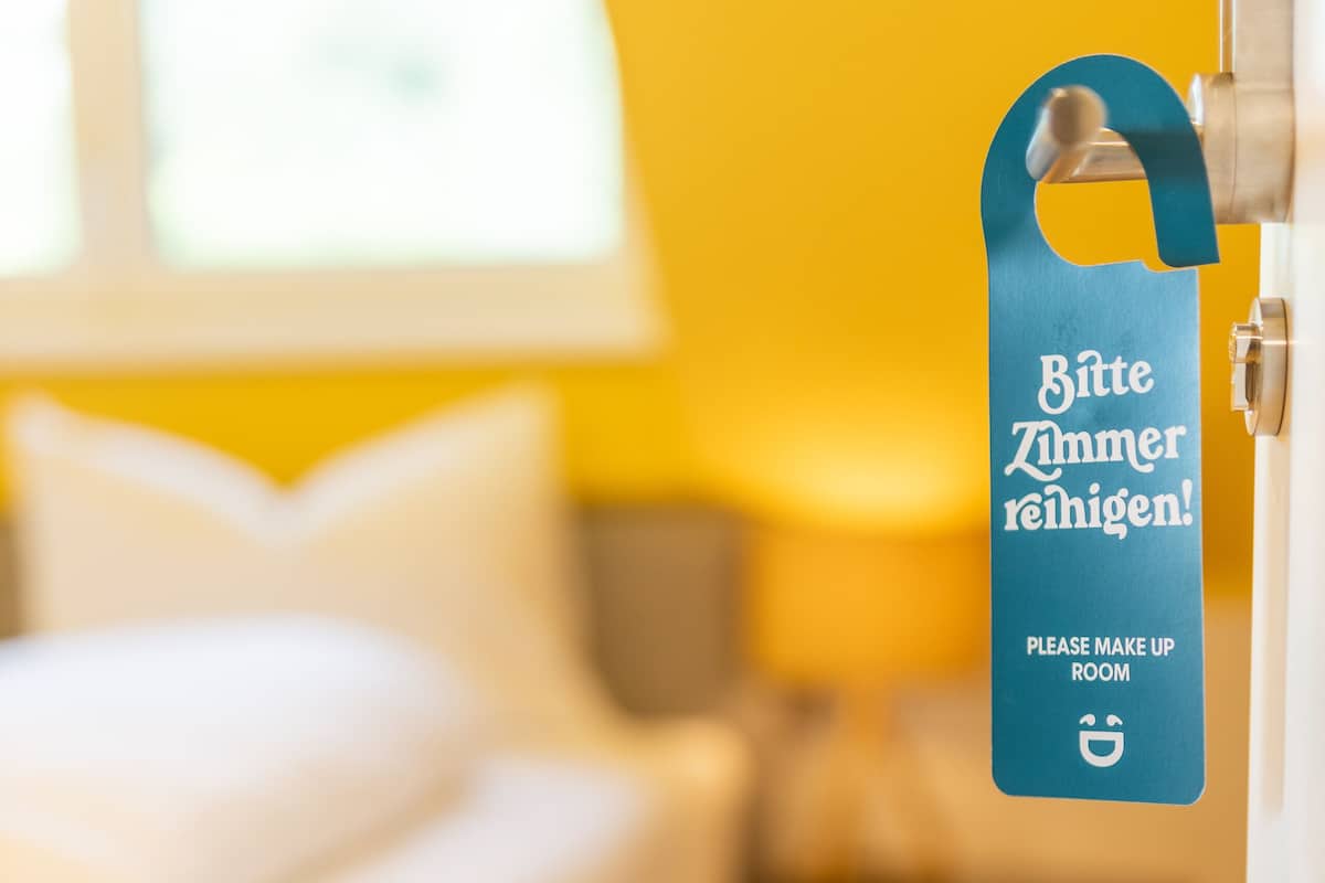 Une chambre avec un lit, un oreiller, une table de chevet et une lampe. Il y a une pancarte sur le mur qui indique : « Veuillez nettoyer la chambre ! »