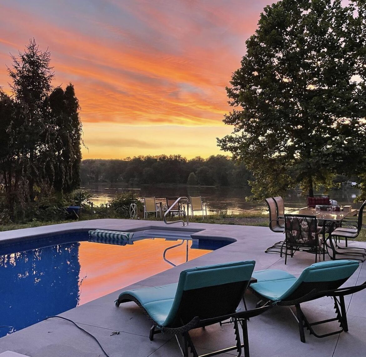 A swimming pool with lounge chairs and a table, surrounded by trees and a view of a body of water during sunset.