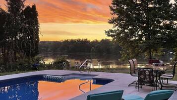 A swimming pool with lounge chairs and a table, surrounded by trees and a view of a body of water during sunset.