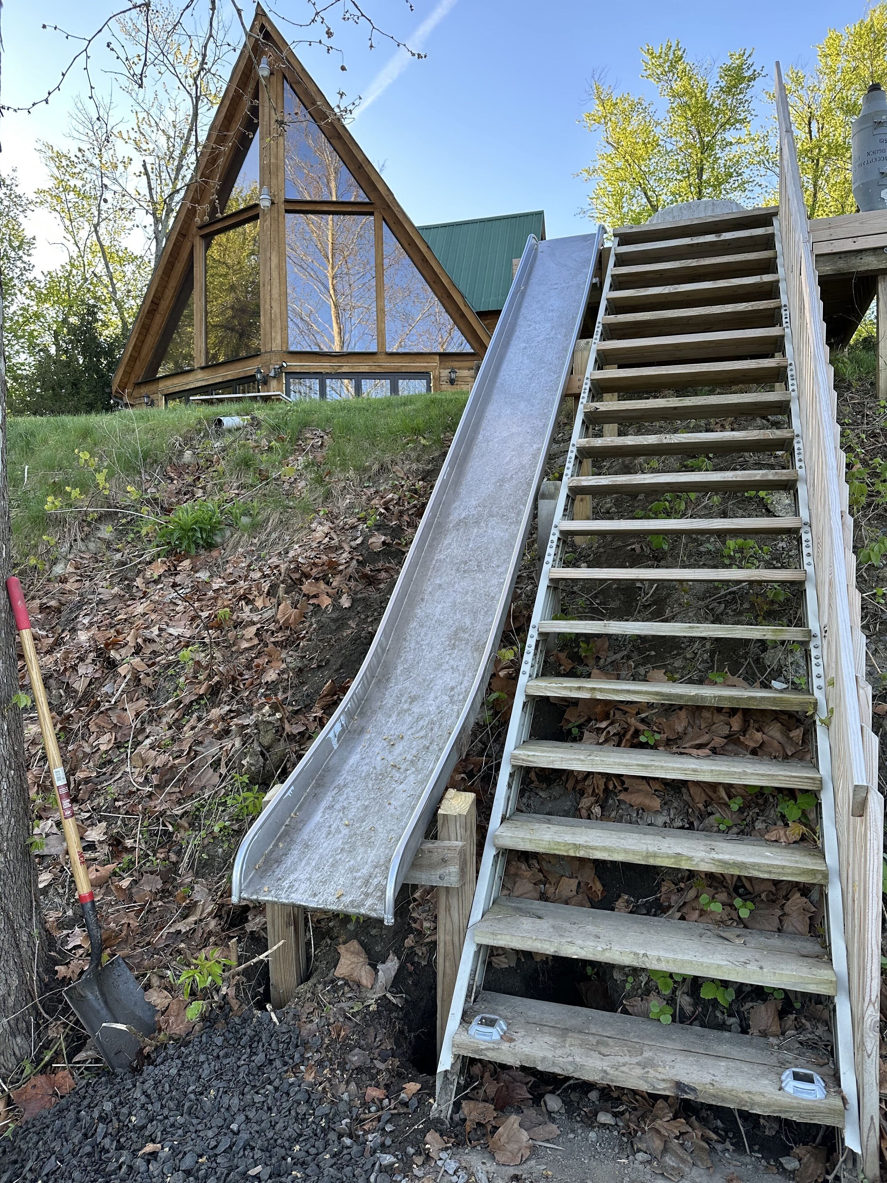A wooden slide attached to a wooden staircase, leading to a building with large windows and a green roof.