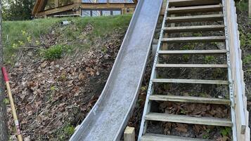 A wooden slide attached to a wooden staircase, leading to a building with large windows and a green roof.