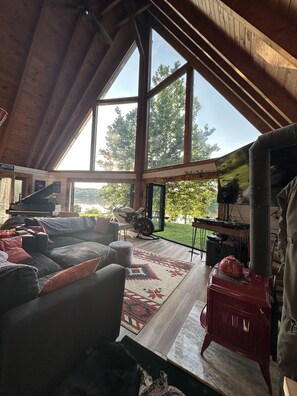 A spacious living room with a large window, a grand piano, a motorcycle, and a wood stove. - Dream A-Frame Cabin on the Hudson with heated pool and dock! Sleeps 10; Pets ok! (Troy)