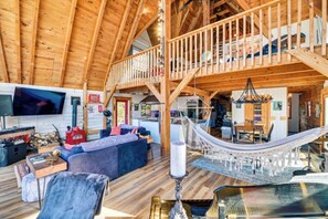 A spacious loft-style living area with a high ceiling, wooden beams, a large sofa, a dining table, and a hammock. - Dream A-Frame Cabin on the Hudson with heated pool and dock! Sleeps 10; Pets ok! (Troy)