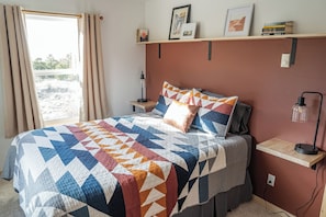 A bedroom with a bed, a nightstand, a lamp, a shelf with books, and a window with curtains.