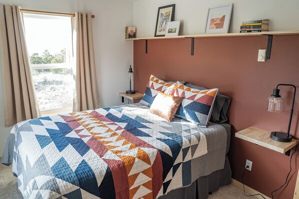 A bedroom with a bed, a nightstand, a lamp, a shelf with books, and a window with curtains.